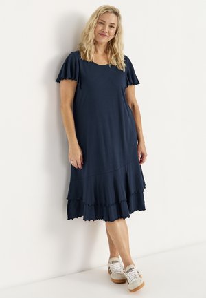 Lyshåret kvinde med bølget hår iført en navyblå lagdelt knælængde kjole og hvide sneakers, stående afslappet mod en hvid væg.