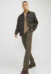 Mann in einer dunkel karierten Jacke mit zwei Brusttaschen, einem beigen Henley-Shirt darunter und karierten Hosen; trägt braune Schuhe.