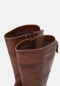 Bottes en cuir marron hautes avec des coutures visibles, doublure en polaire intérieure et fermeture éclair latérale partiellement ouverte sur fond blanc.