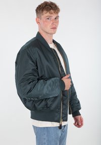 Zelena bomber jakna izrađena od sjajnog, laganog materijala. Ima rebraste manžete, donji rub i zlatni zatvarač s kožnom trakicom.