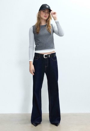 Jeune femme portant une casquette noire, un gilet court gris par-dessus un haut à manches longues gris clair, un jean large foncé et des chaussures noires à bouts pointus.