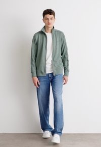 Lysegrønn zip-up jakke over en hvit t-skjorte, kombinert med lyseblå jeans og hvite sneakers. Stilt mot en enkel hvit bakgrunn.