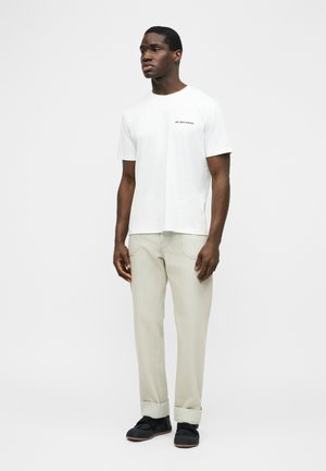 Uomo in piedi che indossa una maglietta bianca a maniche corte con la scritta "Les Benjamins", pantaloni beige con risvolto e scarpe nere su uno sfondo semplice.