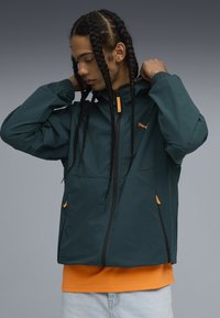 Chaqueta resistente al agua de color teal con capucha, cremallera negra, detalles en naranja y puños elásticos; llevada sobre una camiseta naranja y jeans de color azul claro.