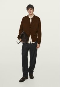 Brauner Cardigan mit Knöpfen, getragen über einem cremefarbenen Hemd; dunkle Hose; hält eine dunkle Ledertasche; glatte Textur mit einem klassischen Schnitt.