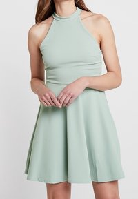 Robe halter vert clair avec un corsage ajusté et une jupe évasée. Fabriquée en tissu lisse, elle présente un col montant et ne comporte aucun motif visible.