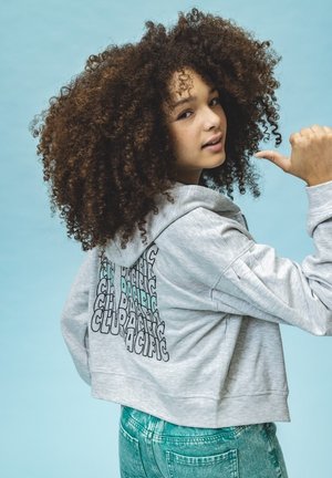 Grijze cropped hoodie met een trekkoord en grafische tekst op de rug in zwart en teal. Gepaard met lichtblauwe spijkerbroek.