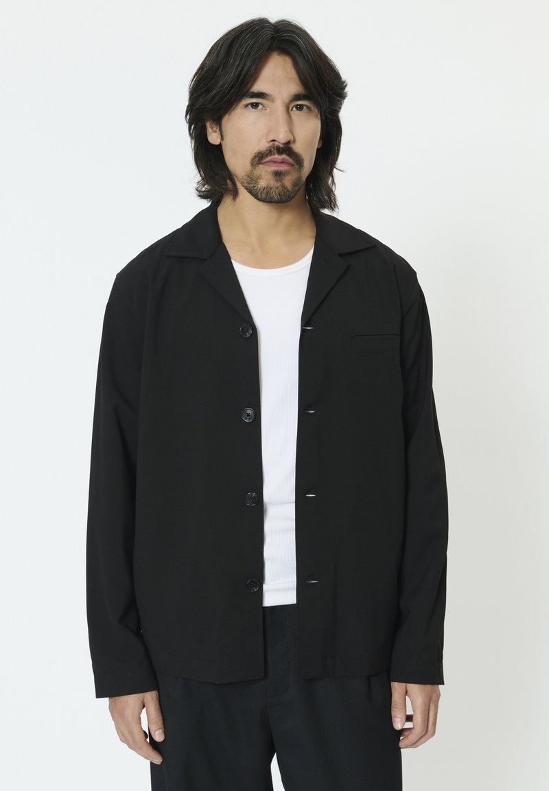 Fekete, gombos dzseki, gallérral, hosszú ujjal és egy mellkasi zsebbel. Sima anyagból készült, laza szabású és finom textúrájú.