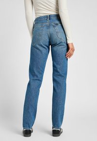 Vaqueros de mezclilla azul con un diseño de pierna recta, con bolsillos traseros y una cintura alta, combinados con zapatos negros y una blusa blanca de manga larga.