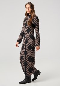 Abito lungo a costine in nero e beige con un motivo geometrico. Presenta un collo alto, un orlo spaccato e bottoni decorativi sulle maniche.