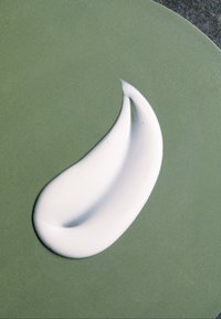 White cream swatch on a textured, muted green surface. The cream has a smooth, glossy finish and a soft curve, with visible product flow marks.