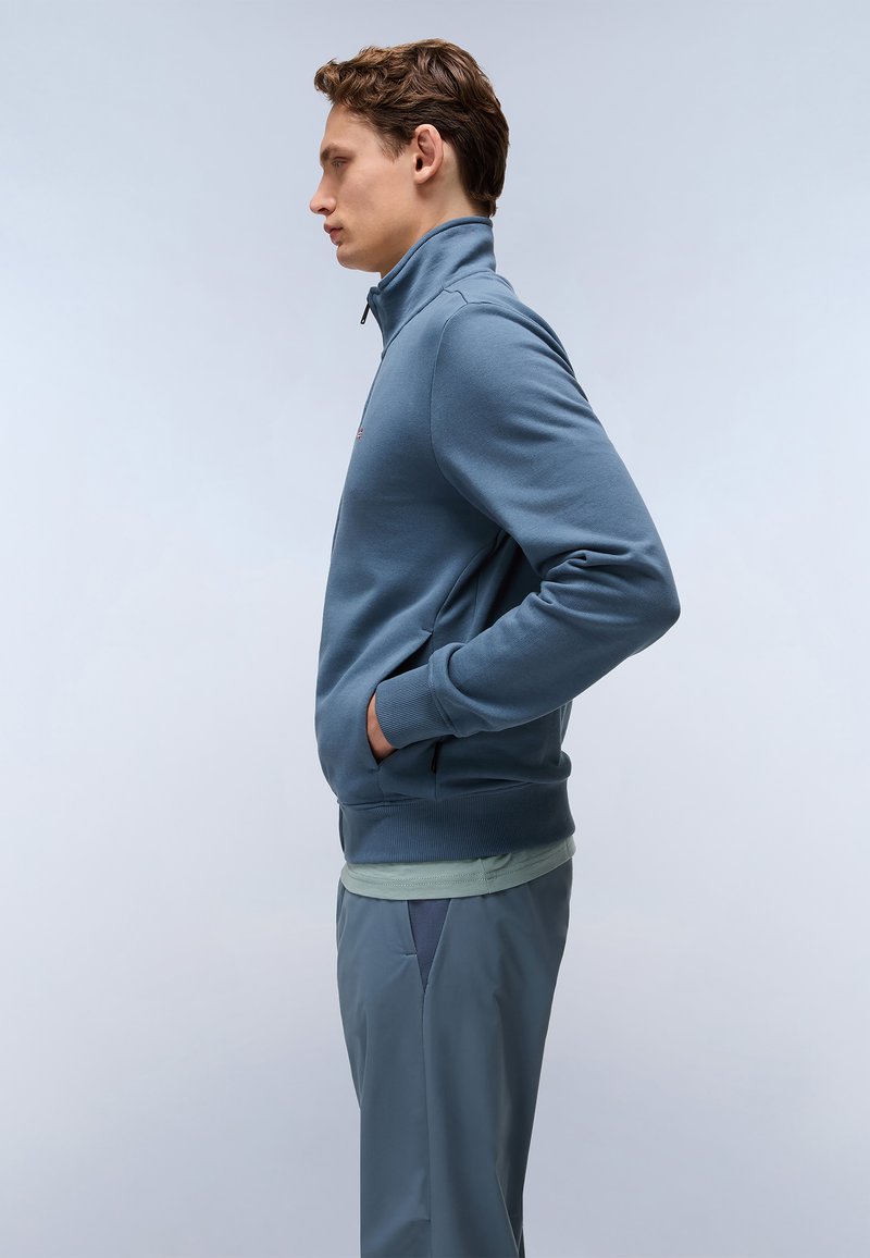 Sudadera azul con cremallera y cuello alto, puños acanalados y bolsillos laterales. Lleva encima una camiseta verde clara, combinada con pantalones gris claro.