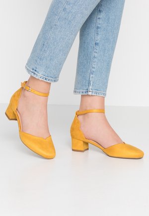 Talons en daim jaune avec un bout arrondi, un petit talon carré et une bride à la cheville. Présente des bords festonnés et une texture lisse.
