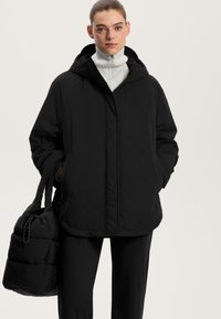 Črna prekrita puhovka s kapuco, ki ima sprednji zadrgo in stranske žepe. Model drži usklajen črn torbico.