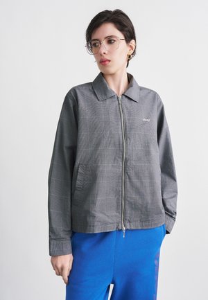 Unge person med kort, mørkt hår, iført klare briller, en grå lynlåstrøje med "obey"-logo og blå bukser, stående foran en hvid baggrund.