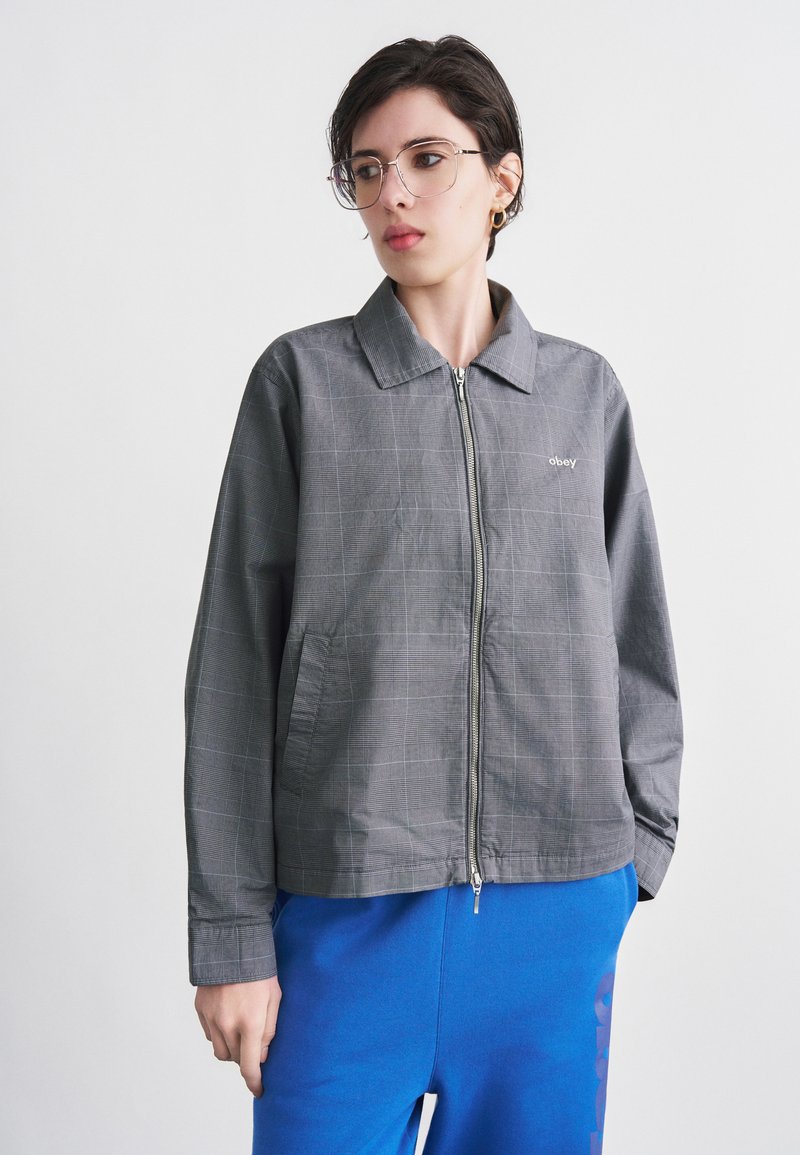 Νεαρό άτομο με κοντά σκούρα μαλλιά, φοράει διαφανή γυαλιά, γκρι ζακέτα με φερμουάρ και λογότυπο "obey", και μπλε παντελόνι, στέκεται μπροστά σε λευκό φόντο.