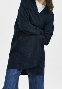 Navy knitted cardigan with a relaxed fit, deep V-neck, long sleeves, and front pockets. Soft texture and low-hem design.