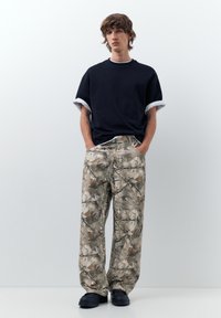 Mladý človek so stredne dlhými vlasmi stojí s rukami v vreckách, oblečený v čiernej rolovanej košeli s krátkym rukávom, širokých nohaviciach s maskovacím vzorom a čiernych topánkach.