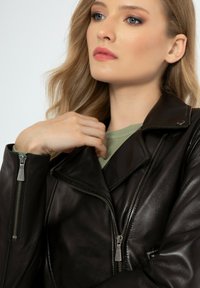 Bruine leren motorjack met zilveren ritsen, waaronder een dubbele rits aan de voorkant, brede kraag en een gladde textuur. Gelaagd over een groene top.