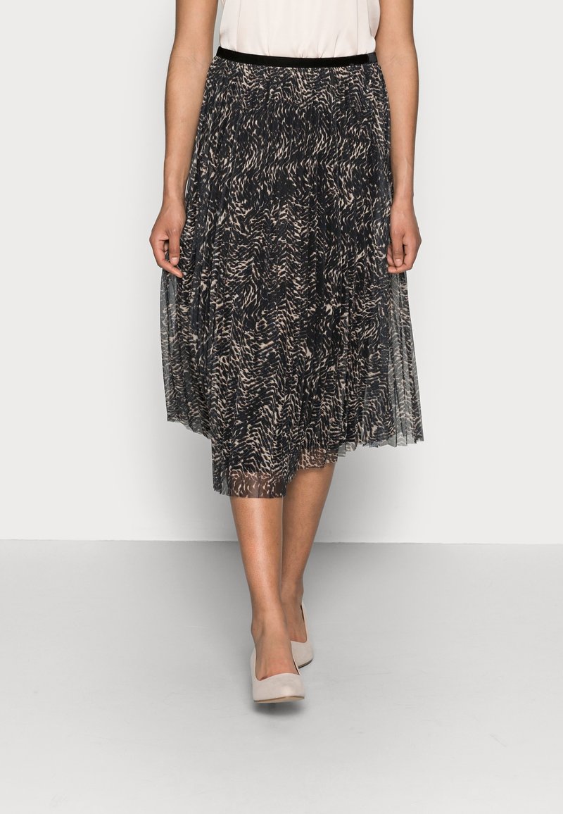 Femme portant une jupe midi plissée noire et beige à motifs, avec un chemisier blanc et des talons beiges, debout devant un fond clair uni.