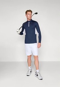 Férfi sötétkék hosszúujjú felsőben, fehér sortban és sportcipőben golfütőt tart a válla felett, egy sima világos háttér előtt áll.