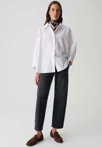 Vrouw staand in een losse witte button-up shirt, zwarte rechte jeans, bruine loafers en een gewaxt halsdoekje op een effen achtergrond.
