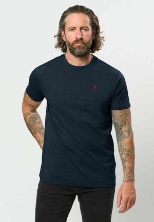 Mand med skulderlangt hår og skæg bærer en navyblå t-shirt med rød logo og viser tatoveringer på begge arme mod en ensfarvet baggrund.