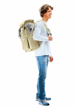 Sac à dos beige avec plusieurs poches et un article en tricot gris attaché, porté par une personne en chemise blanche à manches longues et jeans bleu clair.
