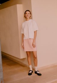 Νεαρή γυναίκα που χαμογελά, στέκεται σε εσωτερικό χώρο μπροστά σε μπεζ τοίχους, φορώντας λευκό φαρδύ t-shirt, ροζ φούστα, λευκές κάλτσες και μαύρα παπούτσια Mary Jane.