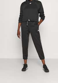 Czarna bluza z kapturem o skróconym kroju, z logo z przodu i bocznymi pasami jako akcentami. Połączona z dopasowanymi czarnymi spodniami dresowymi i butami sportowymi.