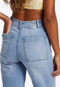 Light blue denim jeans with two large, patch-style back pockets and a label on the waistband, featuring a straight-leg cut.