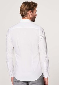 Uomo con capelli castani corti che indossa una camicia bianca a maniche lunghe e pantaloni grigi, visto di spalle su uno sfondo neutro.
