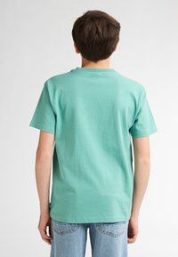 Turquoise T-shirt met korte mouwen, gemaakt van katoen, met een ronde hals. De achterkant heeft een gladde textuur en een rechte zoom. Draagt lichtblauwe jeans.