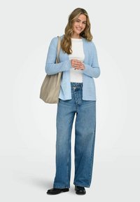 Smiling woman with long hair wearing light blue cardigan, white top, wide-leg jeans, black shoes, and beige shoulder bag on white background.