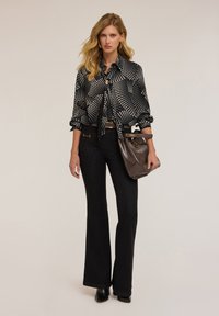 Vrouw in een zwart-wit patronen blouse, zwarte flare jeans, zwarte laarzen, met een bruine leren handtas tegen een beige achtergrond.