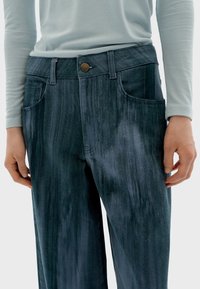 Pantalones oscuros de mezclilla con una superficie texturizada, que presentan un cierre de botón bronce y dos bolsillos frontales. Combinados con una blusa de color azul claro.