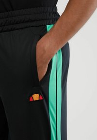 Zwarte sportieve joggers met een brede elastische tailleband, groene zijstrepen en een klein geborduurd logo in rood en oranje.