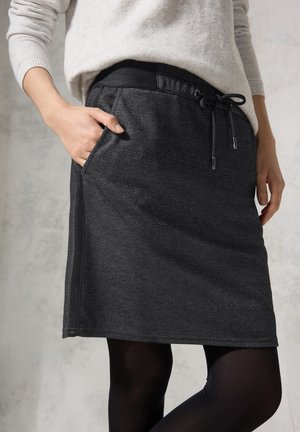Femme portant une jupe trapèze gris foncé avec taille à cordon et poches latérales, associée à des collants noirs et un pull gris clair.