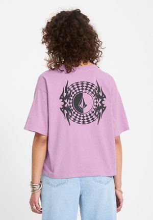 Personne aux cheveux bouclés portant un t-shirt ample lavande avec un motif géométrique circulaire noir et une décoration en forme de flamme dans le dos, associé à un jean bleu clair.