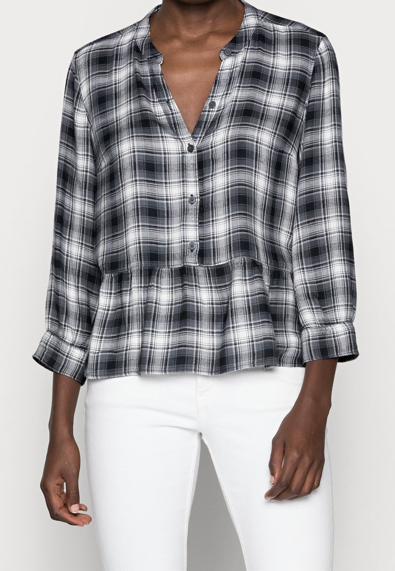 Blusa a cuadros en blanco y negro con botones en la parte delantera, escote en V y dobladillo peplum. Mangas largas con puños abotonados. Textura de tela suave.