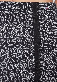 Gros plan sur des leggings noirs avec des motifs abstraits en zigzag blancs et géométriques, une rayure verticale en dentelle noire et un petit logo près de la taille.