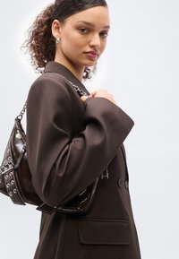 Νέα γυναίκα με σγουρά μαλλιά φοράει ένα σκούρο καφέ σακάκι και κρατά μια γυαλιστερή τσάντα χεριού σε σκούρο καφέ χρώμα στον ώμο της, με φόντο ένα λευκό τοίχο.