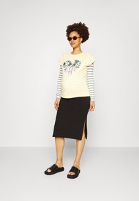 T-shirt amarelo de mangas curtas com gráfico de palmeira, sobre um saiote preto até ao joelho com uma fenda lateral, combinado com sandálias de dedo pretas.