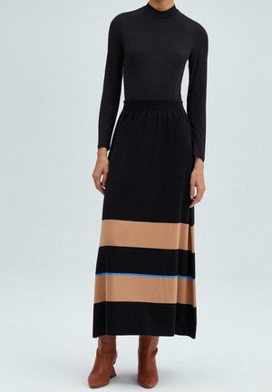 Femme portant un haut noir à manches longues, une jupe maxi noire avec des rayures horizontales beiges et bleues, et des bottines marron.
