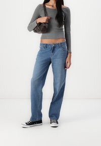 Donna che indossa un crop top grigio a maniche lunghe, jeans blu larghi, sneakers nere, mentre tiene una borsa a tracolla marrone su sfondo bianco.