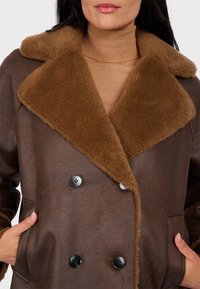 Dwurzędowy płaszcz w ciemno brązowej skórze ekologicznej z dużym brązowym kołnierzem z owczej wełny, z dwoma kieszeniami i czarnymi guzikami.