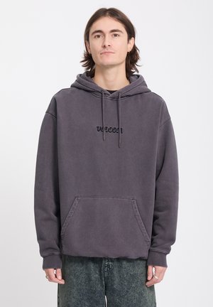 Jeune homme portant un sweat à capuche gris foncé avec poche avant et un jean vert délavé, debout devant un fond blanc uni.