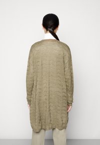 Olivgrüner, gestrickter Cardigan mit lockerer Passform, strukturiertem Zickzackmuster, langen Ärmeln und abgerundetem Saum, getragen über beigen Hosen.