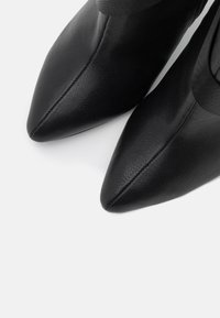 Botas negras de punta afilada hechas de cuero. Presentan una textura suave y costuras visibles a lo largo de las uniones. Diseño minimalista, sin adornos.