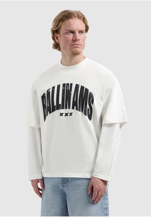 Jonge man met blond haar, gekleed in een witte, gelaagde lange mouw en T-shirt met de tekst "BALLINAMS XXX", lichtblauwe jeans, kijkt opzij.